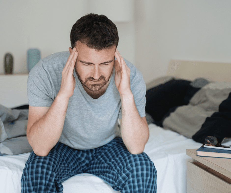 man zit op het randje van zijn bed. Handen zorgelijk tegen het hoofd.