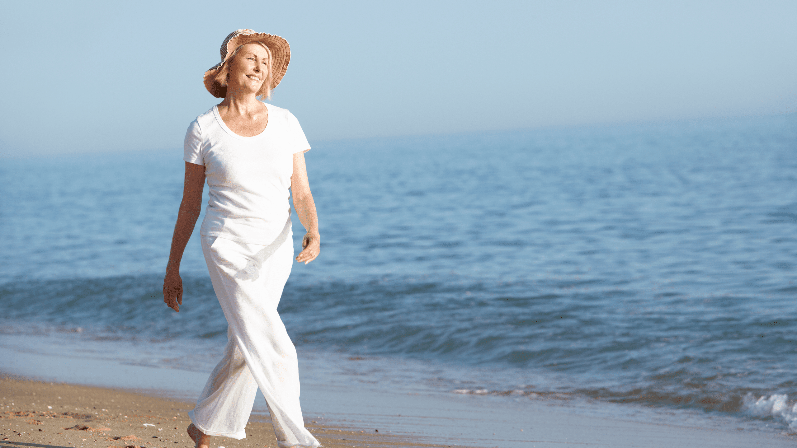 Vitale oudere vrouw wandelt energiek over het strand, langs de kustlijn. Ze is gekleed in witte broek, een wit t-shirt en draagt een zonnehoed.